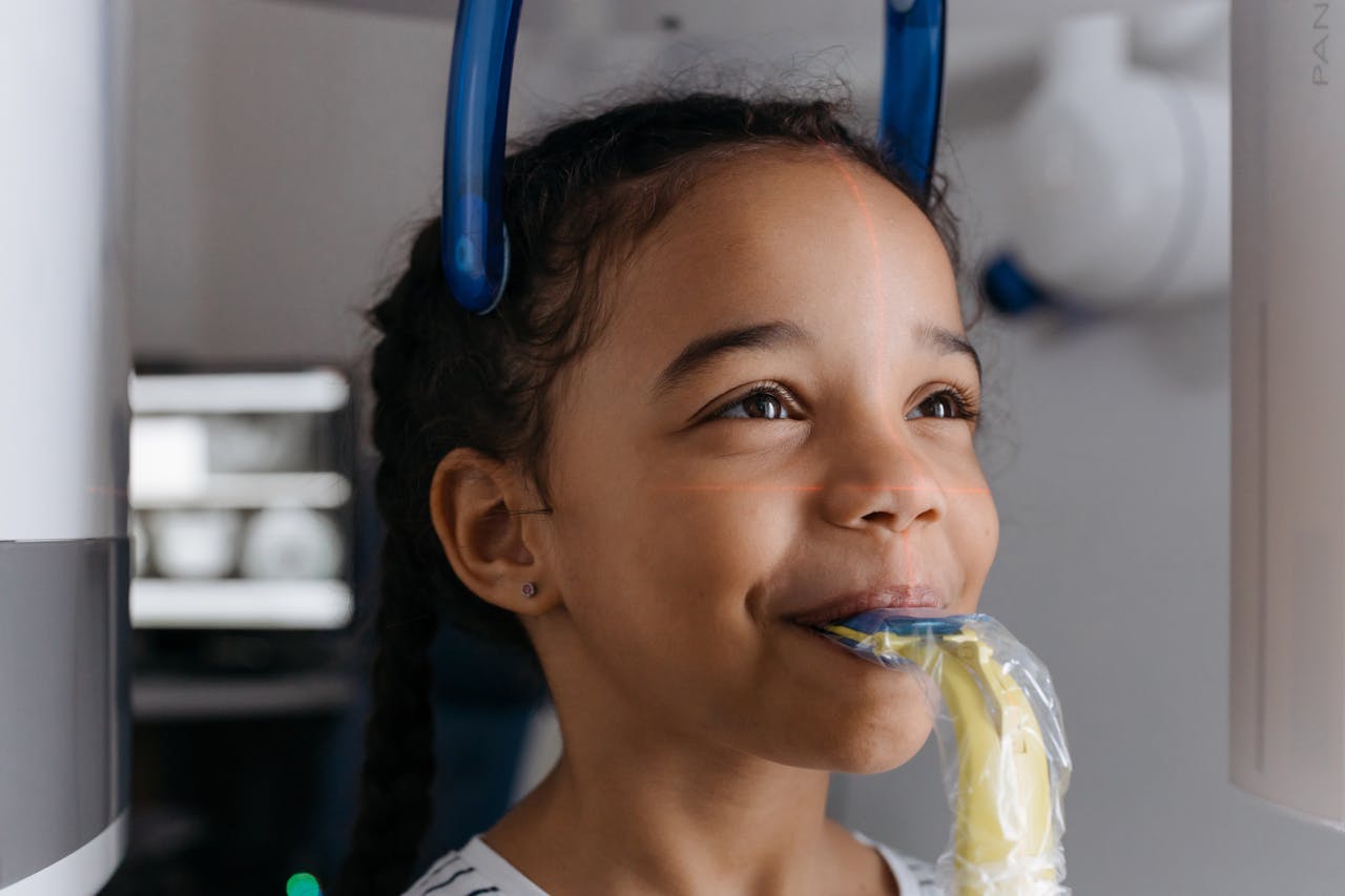 A young girl smiling while undergoing a dental X-ray in a clinic setting.